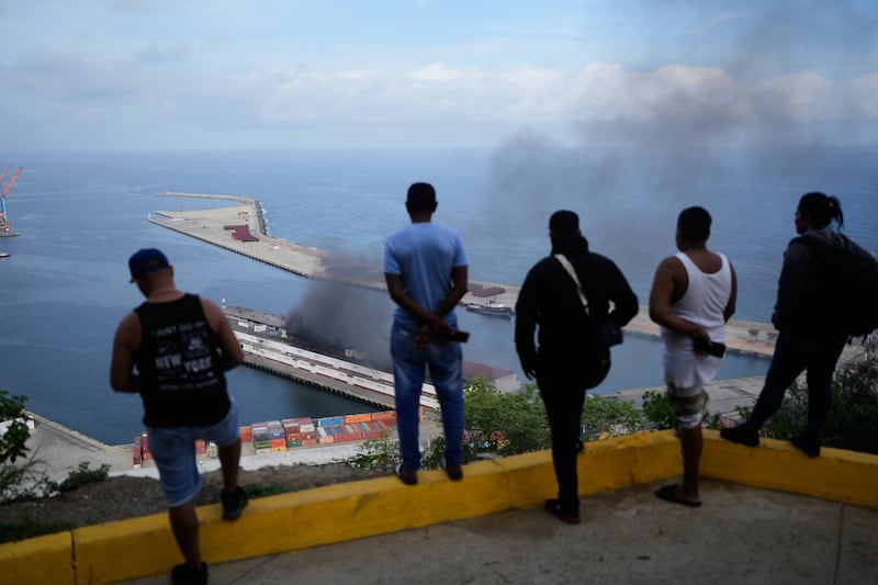 Men watch smoke rising from a dock after explosions were heard at La Guaira port, Venezuela, Saturday, Jan. 3, 2026. (AP Photo/Matias Delacroix)