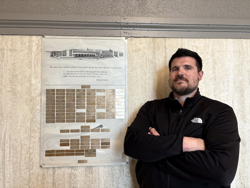 York Community High School history teacher Justin Riskus leads a capital campaign for an updated memorial to the high school's graduates who have died in battle. He stands next to the original memorial plaque donated by York's Class of 1985.