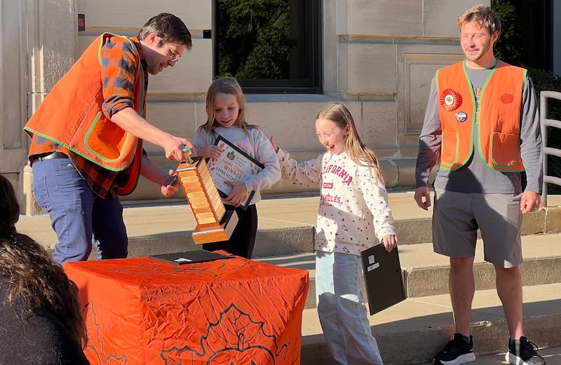 Greg D’Addario shows Rayna Hochstatter on May 20, 2026, the plaque, with her name and winning entry, that has been added to the Sycamore Pumpkin Festival theme contest trophy, while Annabelle Hochstatter and Trenton DeVito watch.