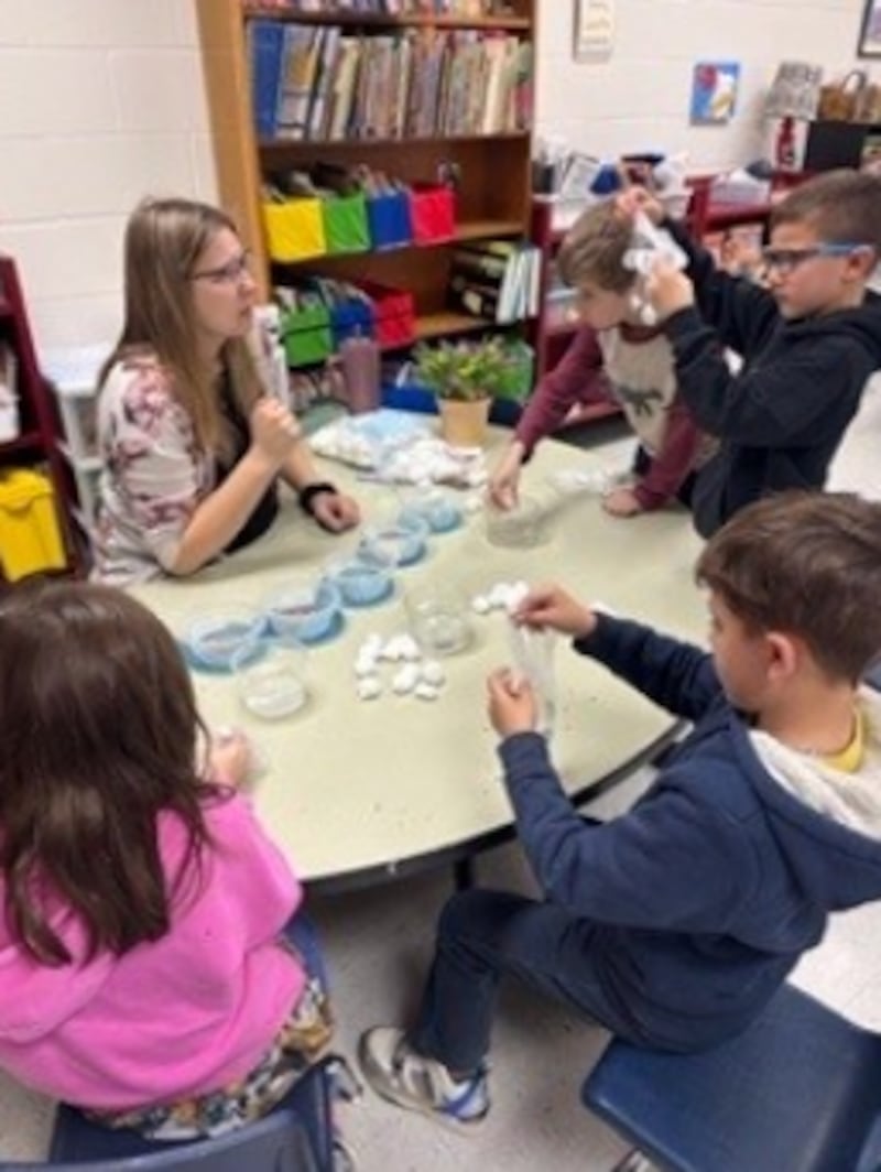Extension Ag in the Classroom Assistant Natalie Blum helps West Carroll first graders wet a cotton ball to add to each finger of the glove to start the geminating process.