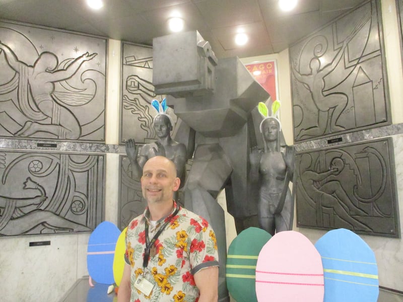 Joliet Central High School teacher Jeffrey Grimes poses for a photo at the Steelman sculpture inside the school. The sculpture setting at the time was decorated for the Easter holiday, and Grimes was dressed for a Hawaiian theme day at the school. April 11, 2025