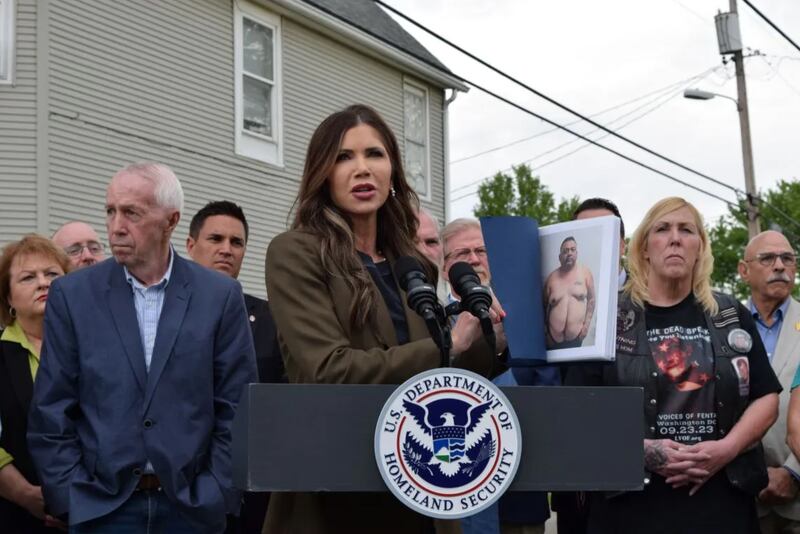 DHS Secretary Kristi Noem speaks in front of a house in the neighborhood where progressive activist Emma Shafer was murdered in 2023. Noem holds a binder full of pictures of people she said are “still at large” after engaging in criminal activity in Illinois.