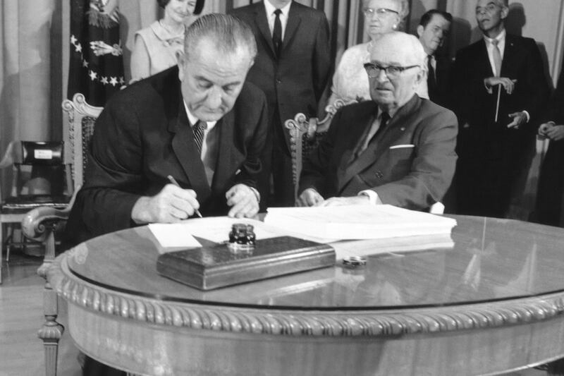 FILE - President Lyndon B. Johnson uses the last of many pens to complete the signing of the Medicare Bill into law at the Truman Library in Independence, Missouri, July 30, 1965, with former President Harry S. Truman at his side. At rear are Lady Bird Johnson, Vice President Hubert Humphrey, and former first lady Bess Truman. (AP Photo, File)