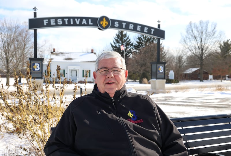 Former longtime Bourbonnais Mayor Paul Schore sits along Festival Street, part of the village's recent park development, The Grove, and across from the Bourbonnais Grove Historical Society and the 1837 Log Schoolhouse Restoration project, at right. Schore was instrumental in the completion of both historical projects.