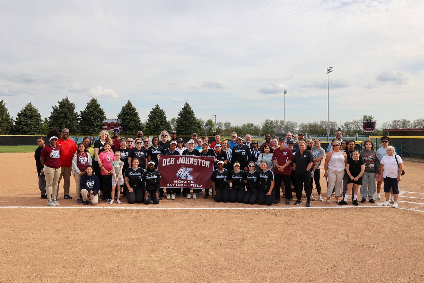 Kankakee High School players, staff, family and friends of the late Deb "Coach. J" Johnston pose for a photo with the new banner designating the softball field as the Deb Johnston Memorial Softball Field on Wednesday, May 7, 2025.