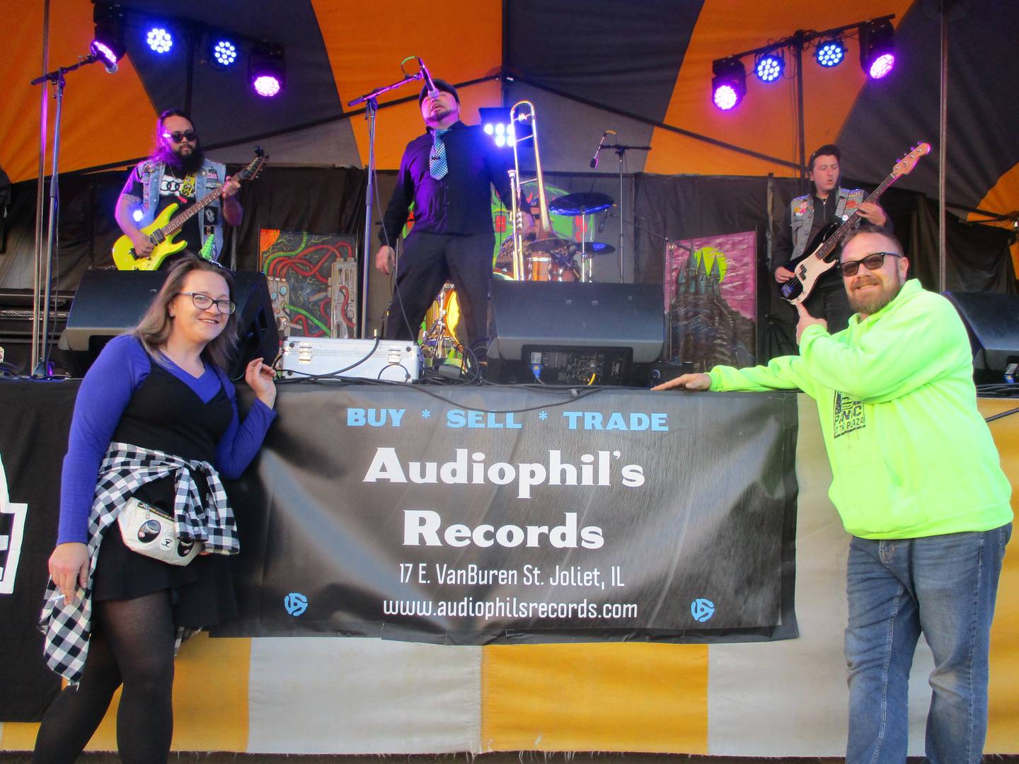 Tiffany and Phil Johnson pose in front of a band performing on the stage for Panic at the Plaza, the event they organize each year and which was held Saturday, Oct. 7, 2023 in downtown Joliet.