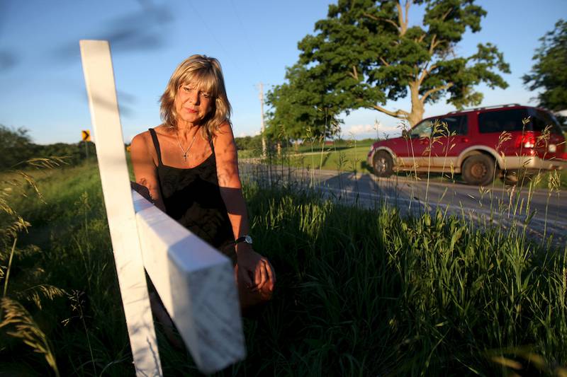 Vicki Moore kneels down June 20 next to a cross for her son as cars drive around the sharp turn of Plank Road near Moose Range Road in Sycamore. Jonathan Moore, Vicki’s son, was killed there as a passenger in a drunken driving crash in the early morning of June 21, 2003.