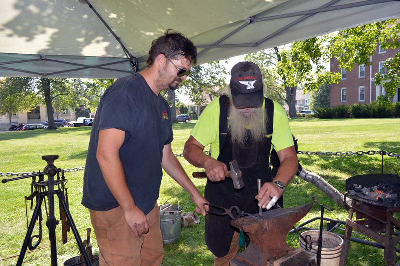 Lloyd Bellows (right), of Pine Creek Forge, engraves a design on a spoon handle while Matt Modrzejewski uses tongs to hold the burning hot metal Saturday, Aug. 24, 2024, during StrawFest in downtown Mt. Morris. StrawFest was a part of the ninth annual U.S. National Straw Sculpting Competition.