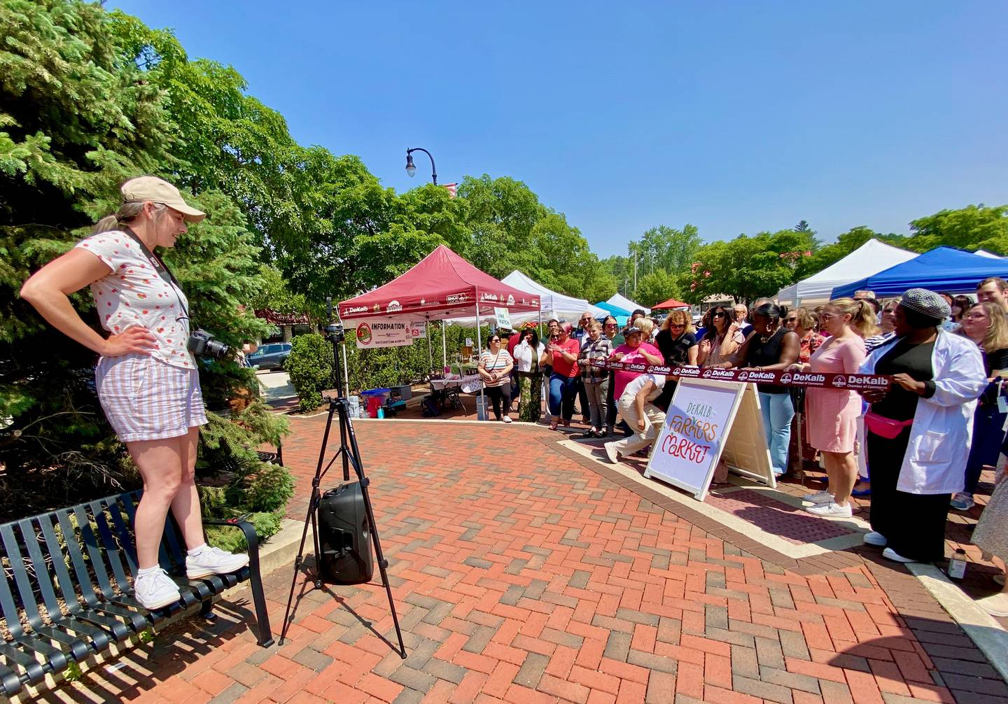 DeKalb Chamber of Commerce membership manager Anna Wilson (left) readies the crowd ahead of a ribbon cutting to celebrate the opening day of the DeKalb farmers market, on Thursday, June 5, 2025, in Van Buer Plaza in downtown DeKalb. The market opened its 30th year Thursday.