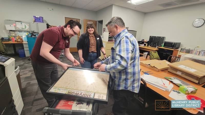 Adult Services Manager Zach Terrill, Youth Services Librarian Lead Steffanie Baseley and Building Services Manager Michael Schnaebele (left to right) install the new StoryWalk in Knox Park in McHenry.