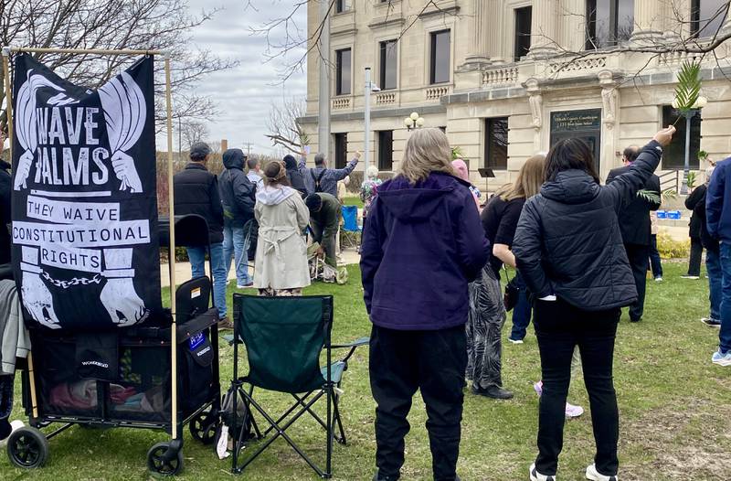 A sign that says, "We wave palms. They waive constitutional rights," is posted next to worship service goers during a Palm Sunday Faith Action event on Sunday, March 29, 2026, in front of the DeKalb County Courthouse in Sycamore. Area Christian ministers organized the event to combat the rise of Christian nationalism in the U.S. Those gathered held palm branches as they sand hymns.