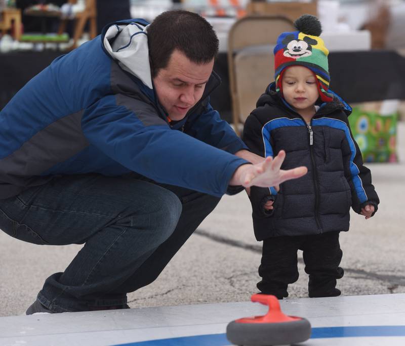 Chris Lalla of Oak Brook demonstrates curling for his son, Mason, 1, during Frozen Fest in Glen Ellyn Saturday.